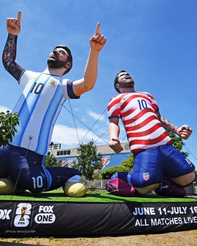 Two large inflatable soccer players kneeling, one in an Argentina jersey, the other in a USA jersey, with a giant ball nearby. Clear blue sky backdrop.