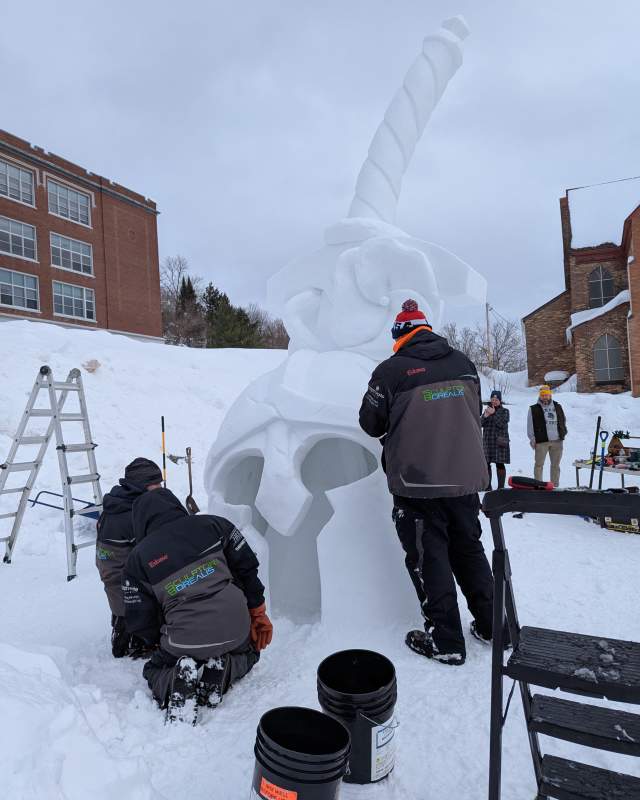 three people put finishing touches on a snow sculpture of a roman helmet pierced by a sword