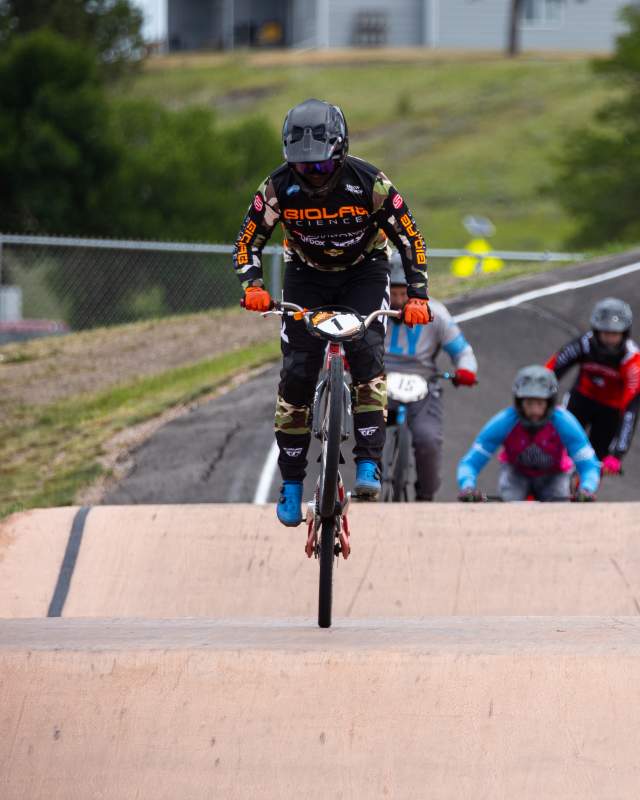 Racing on a paved BMX track, a cyclist in a black racing jersey and helmet navigates a jump. Four other racers are behind him in the background.
