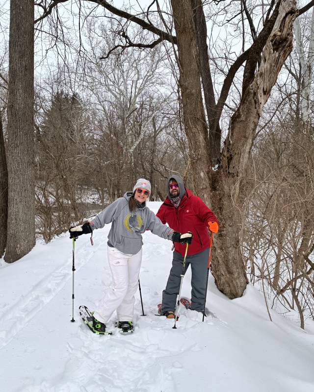 Two people smiling while snowshoeing in the snow. One person is wearing a gray sweatshirt and the other is wearing a red jacket. There are trees in the background.