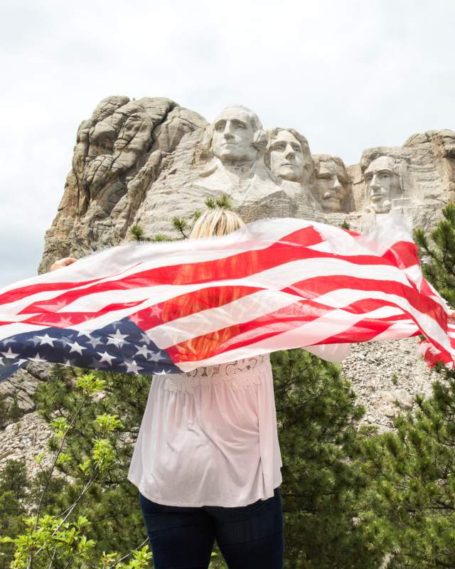A woman holding an American flag behind her faces away from the camera toward Mount Rushmore, visible in the upper half of the photo under a mostly cloudy sky.