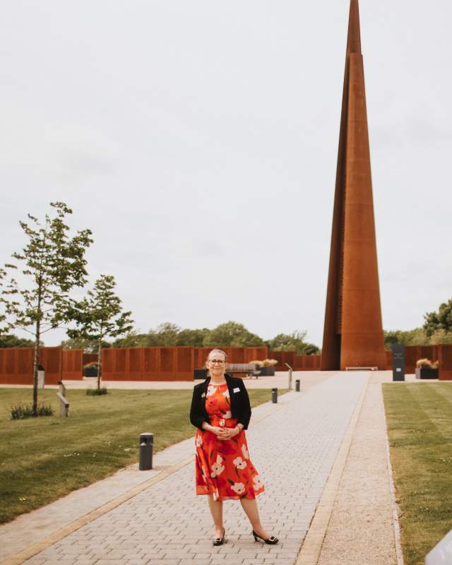 a woman stood on a path surrounded by fields and trees with a spire monument in the background