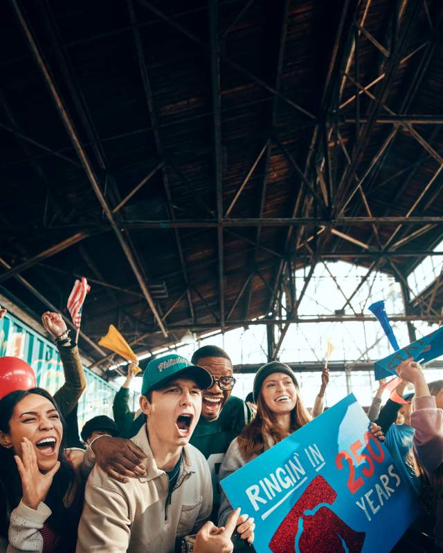 A group of people cheering in a warehouse with a sign that reads Ringin in 250 Years