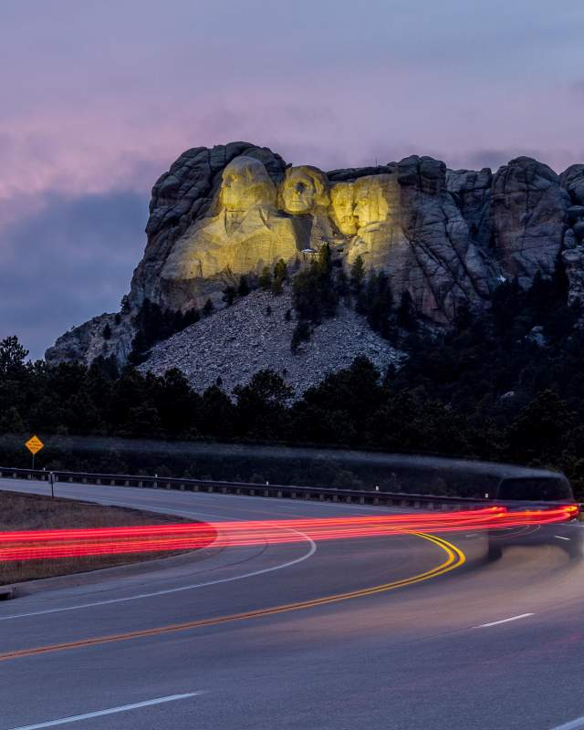 Mount Rushmore, lit up by yellow floodlights at dusk, sits in the background as a vehicle traveling down the highway is trailed by a long line of red taillights, created by a long-exposure photo.