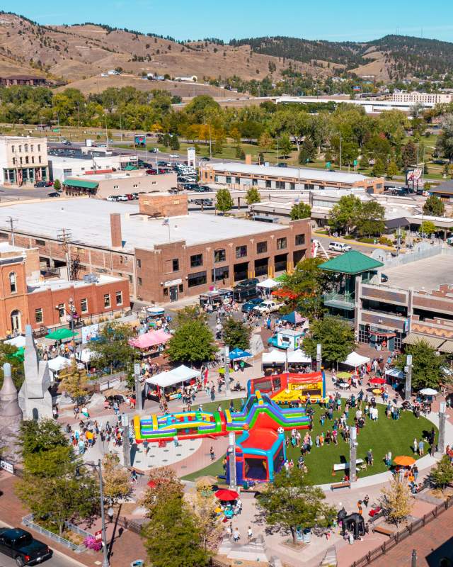 Aerial view of a busy festival at Main Street Square in downtown Rapid City, with crowds gathered around colorful inflatable play structures, vendor tents, and a green event lawn, surrounded by historic brick buildings and parking garages, with the rolling hills of the Black Hills in the background.