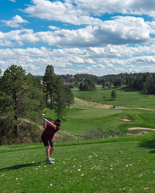A man dressed in a black polo and black shorts tees off at the Golf Club at Red Rocks. Pine trees, a perfectly manicured course, bunkers, and a partly cloudy sky are visible in the distance.