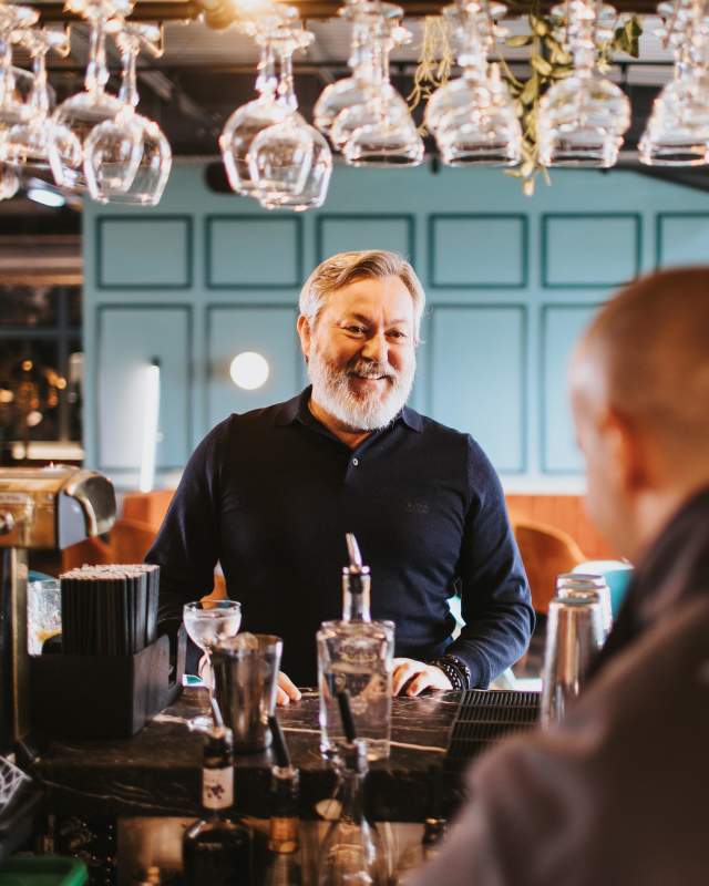 a man looking at bar staff over a bar with glasses hanging from a rack