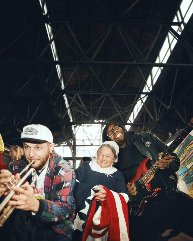 Three people smile and perform music inside a large industrial-style building—one playing trumpet, one playing electric guitar, and a woman in historical dress holding a flag.