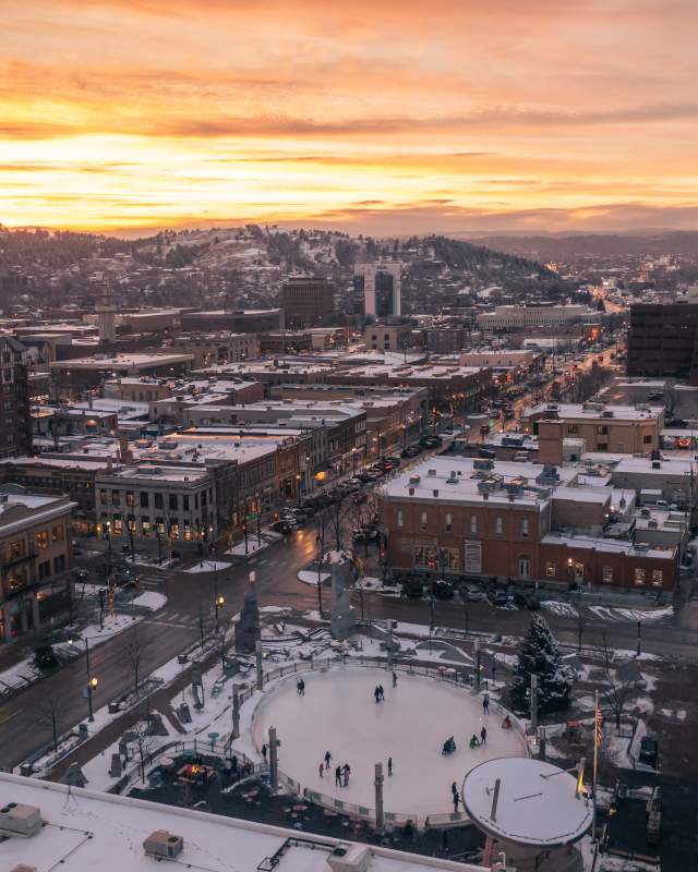 A snowy cityscape at sunset, with warmly lit buildings and streets. A central ice rink with skaters is surrounded by hills and a vibrant orange sky.