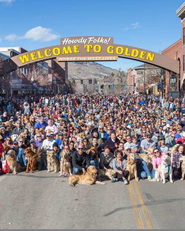 Golden retrievers and owners gather under "Welcome to Golden" Arch
