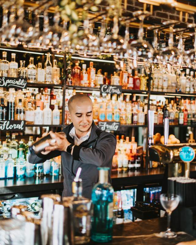A barman shakes a cocktail behind a bar, as a customer patiently waits for his drink.