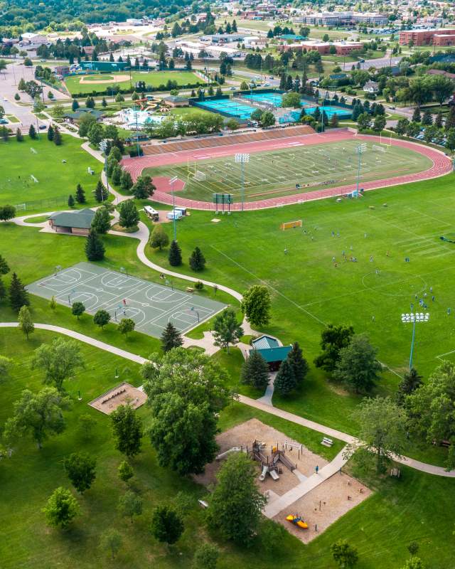 Birds eye view of soccer field, playground, horseshoe, track, basketball court, and pool surrounded by housing.