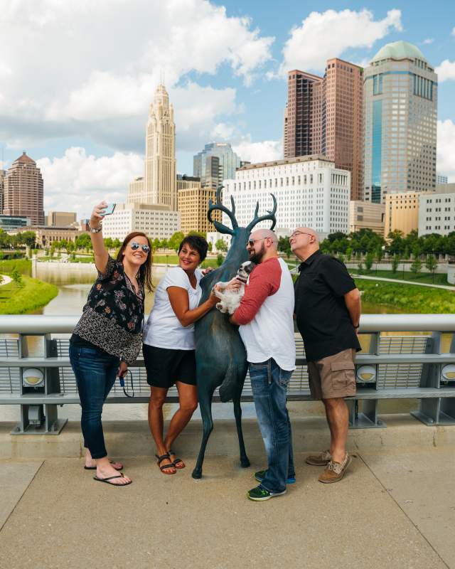 Friends taking picture with deer statue along Scioto Mile. Downtown skyline in the background.