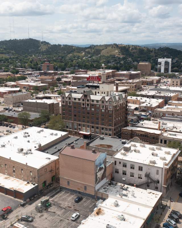 An aerial shot of downtown Rapid City, with the Block 5 building and the Hotel Alex Johnson prominently in the foreground, Skyline Drive slightly out of focus, and the silhouette of the Black Hills on the horizon.