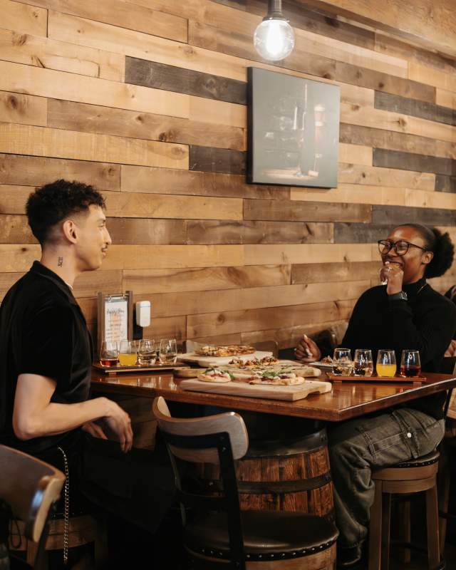 One man and one woman, both dressed in black shirts, sit across a table from each other at Bin605. Their table has an assortment of food items as well as a four-glass wine flight. The restaurant is well lit by modern light fixtures and is accented by a wood palate wall behind them. Other restaurant patrons sit at a table behind the pair, falling off of the right side of the photo.