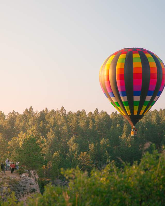 Colorful hot air balloon rising over the Black Hills National Forest with spectators watching cliffside.