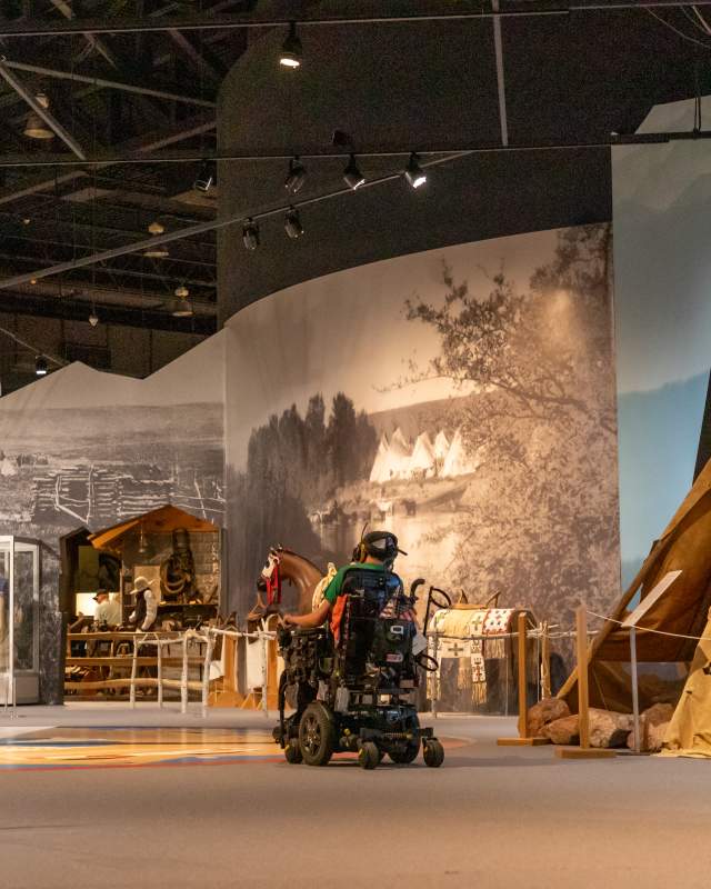 A man in a wheelchair makes his way through the Lakota history section of the Journey Museum, with two replica teepees on either side of a large walk-through exhibit.