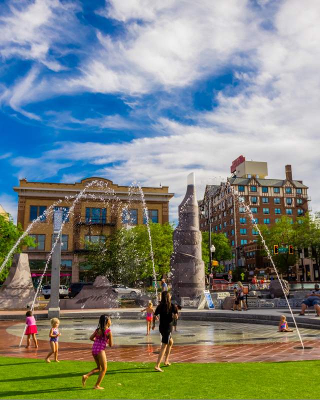 Children running through a splash patio next to a patch of green and surrounded by charming buildings. Blue sky with clouds.