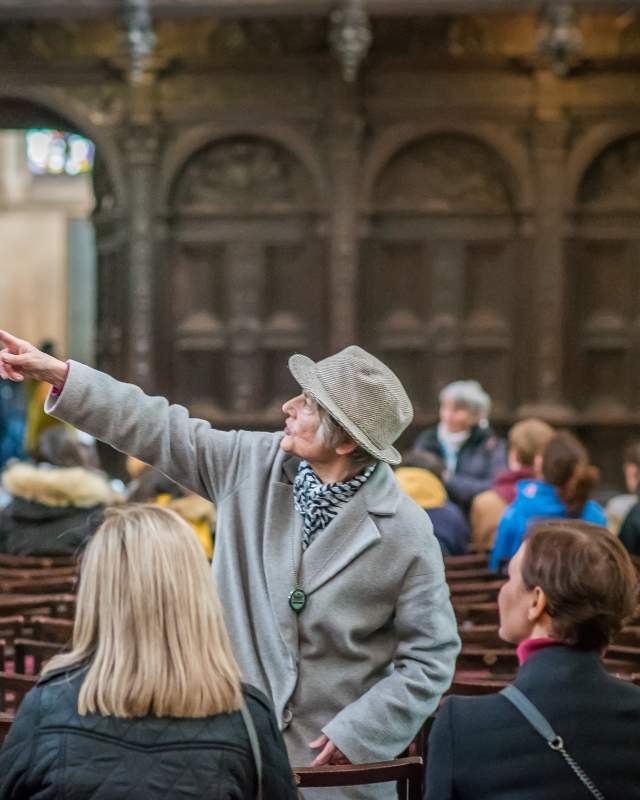 White haired white woman in a grey wool coat & grey felt hat, standing profile pointing to the left in Kings College Cambridge Chapelpointing