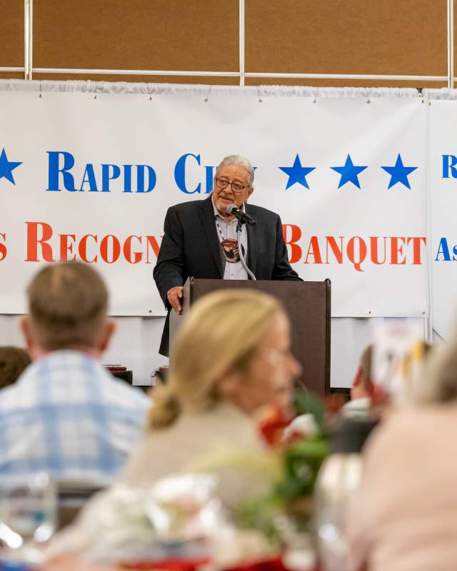 A man gives a speech at a podium on a stage in front of a banner that reads "Rapid City Sports Recognition Banquet" as guests look on from tables on the floor.