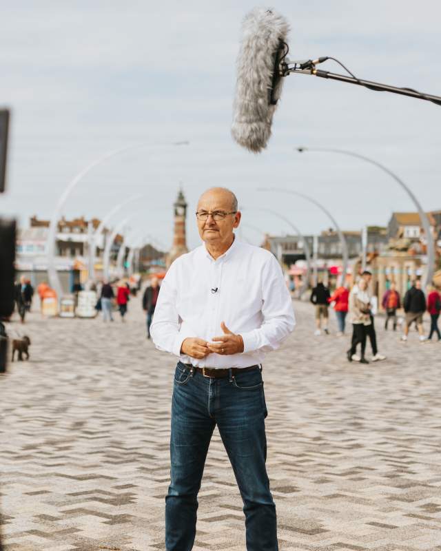 A man stood on skegness promenade with a clock tower in the background and a camera crew out of focus in the foreground