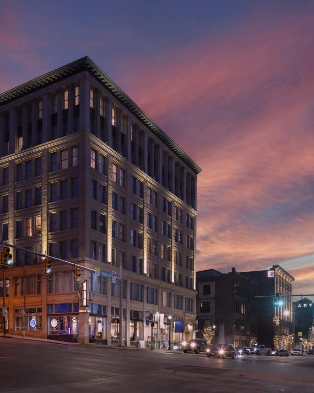 The illuminated Hotel BLU, a historic downtown building, stands on a city street corner beneath a vibrant pink and purple sunset sky.