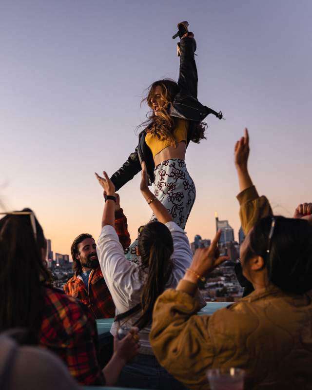 A woman dances with a crowd on a rooftop, overlooking a city skyline.