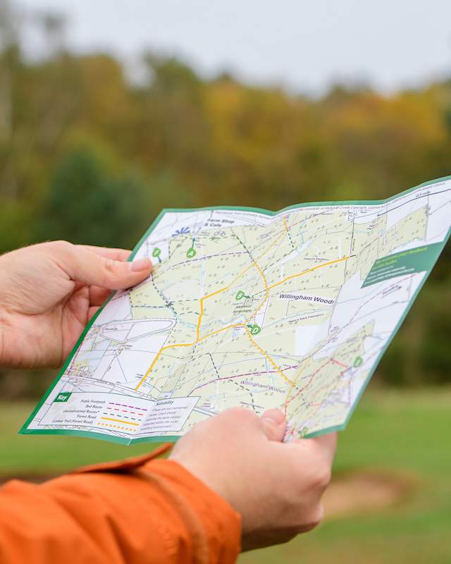 a close up of a man holding a walking guide map with a bright orange coat on and autumnal trees in background