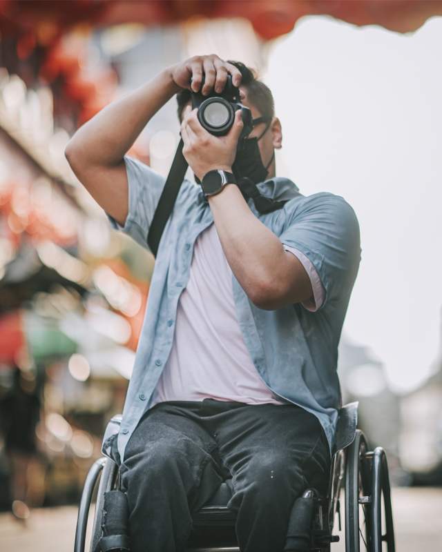 A man in a blue shirt takes photos with a camera while sitting in a wheelchair.
