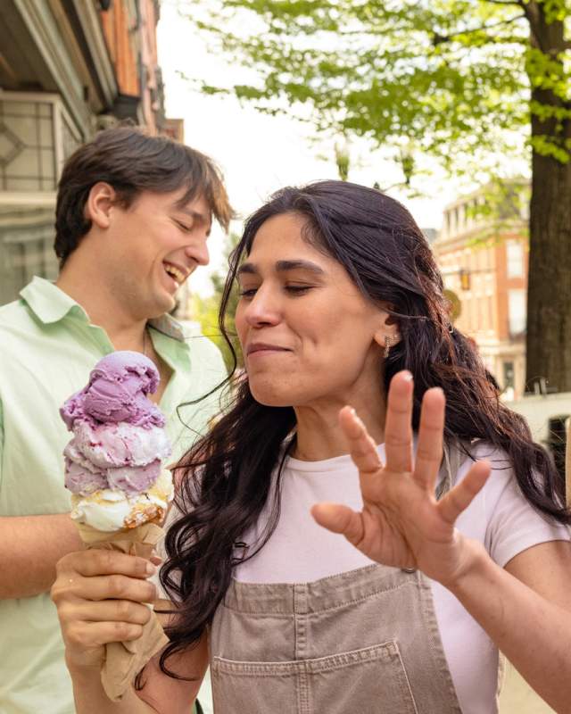 A woman with a large ice cream cone turning away from another woman laughing with a milkshake in her one hand and running her other hand through her hair on a sidewalk on a main street. Two men are giggling and standing around them