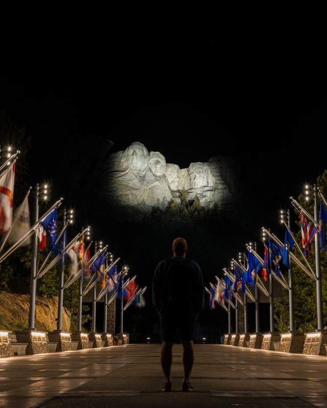 person standing at the avenue of flags looking up at an illuminated mount rushmore in the black hills of south dakota