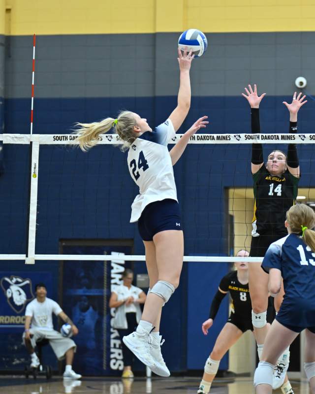 A volleyball player in a white jersey leaps to tap the ball over the net above two defenders in black jerseys.