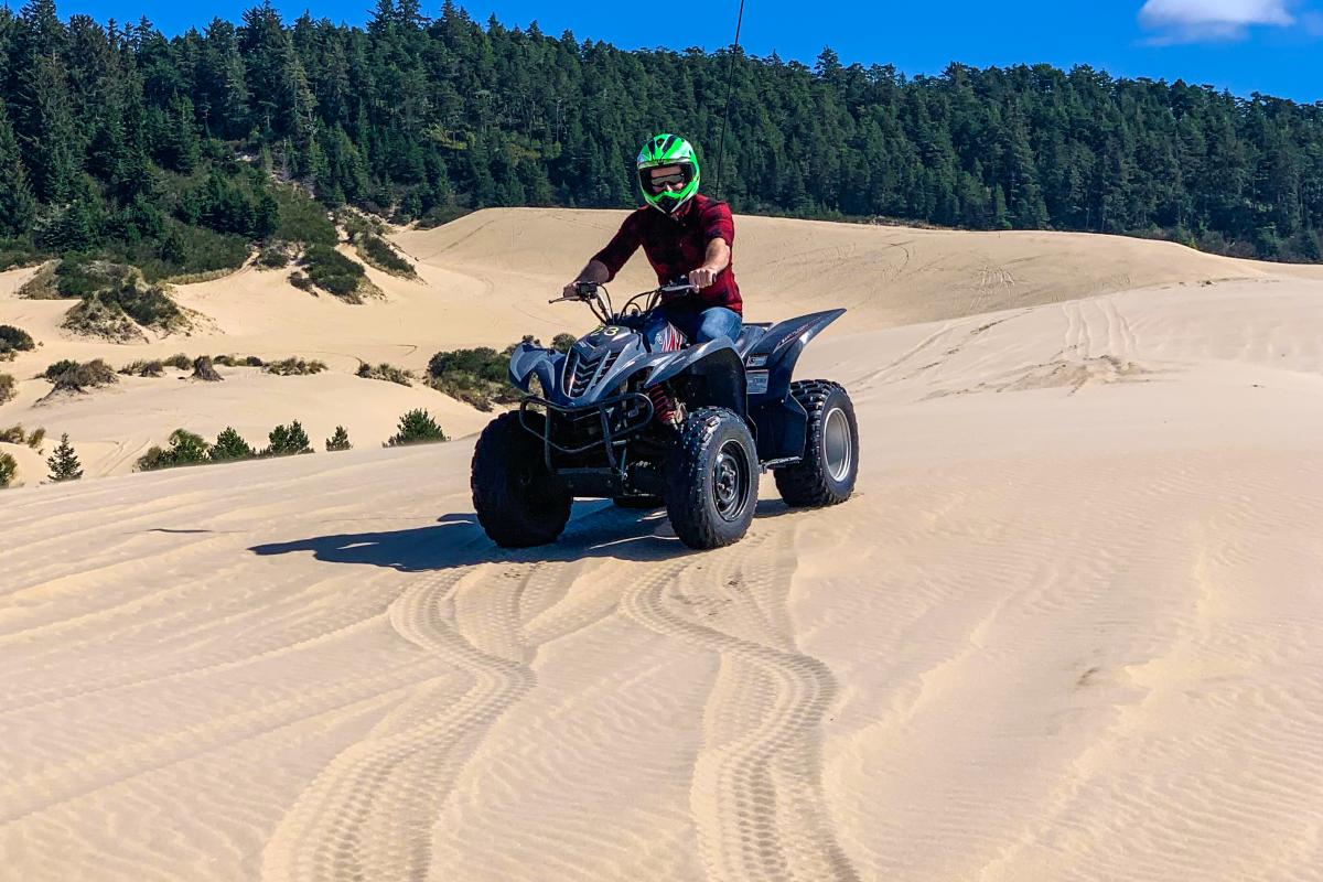 A man sits on an ATV on top of a large sand dune with forest in the background.