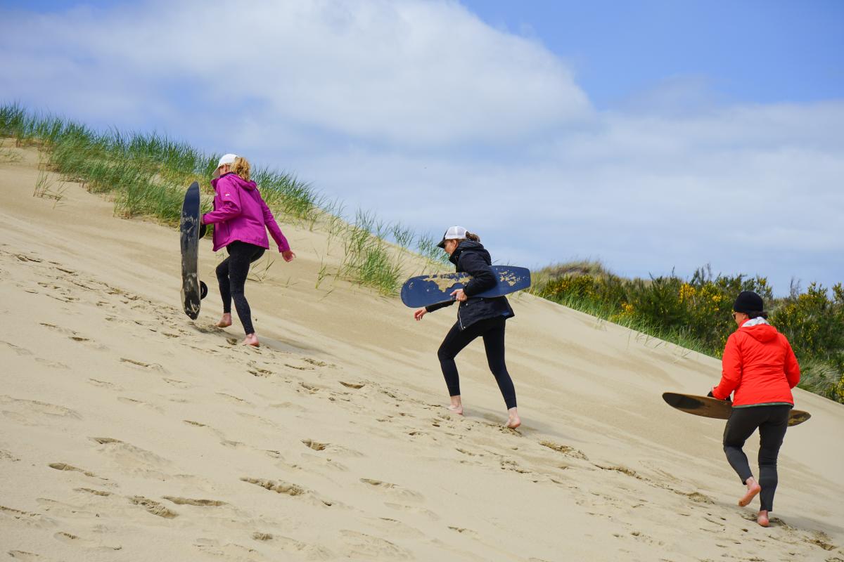 Three women with sandboards hike up a dune.