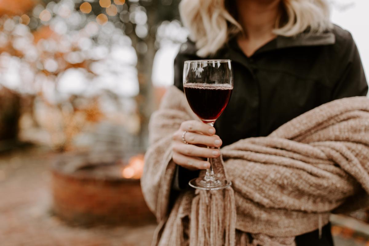 A hand holds a glass of red wine. There is a shawl wrapped around the arms of the person holding the wine glass. Fall foliage is in the background.