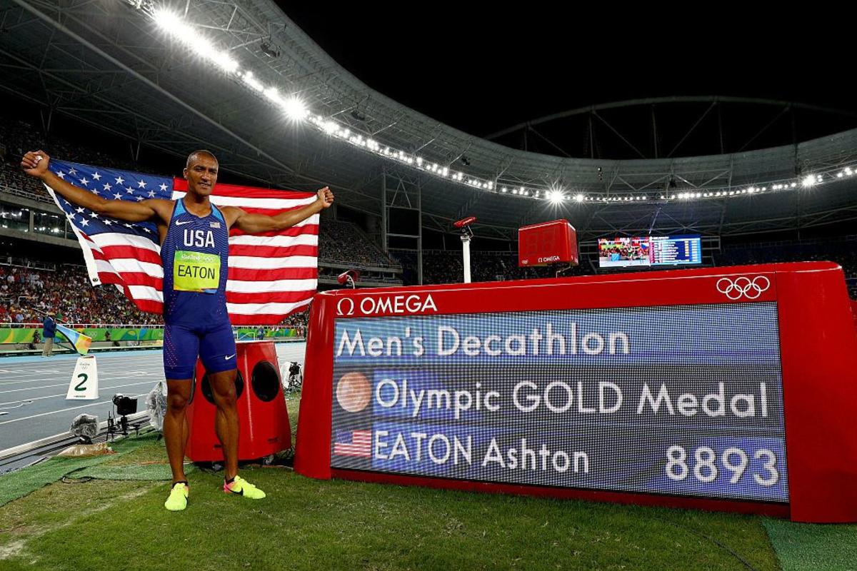 Ashton Eaton stands with an American flag wrapped around him. Beside him is a display that says Men's Decathlon Olympic Gold Medal