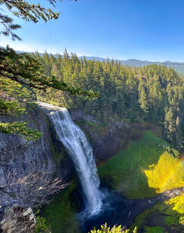 A view of a large waterfall in a vast forested canyon on a sunny day.