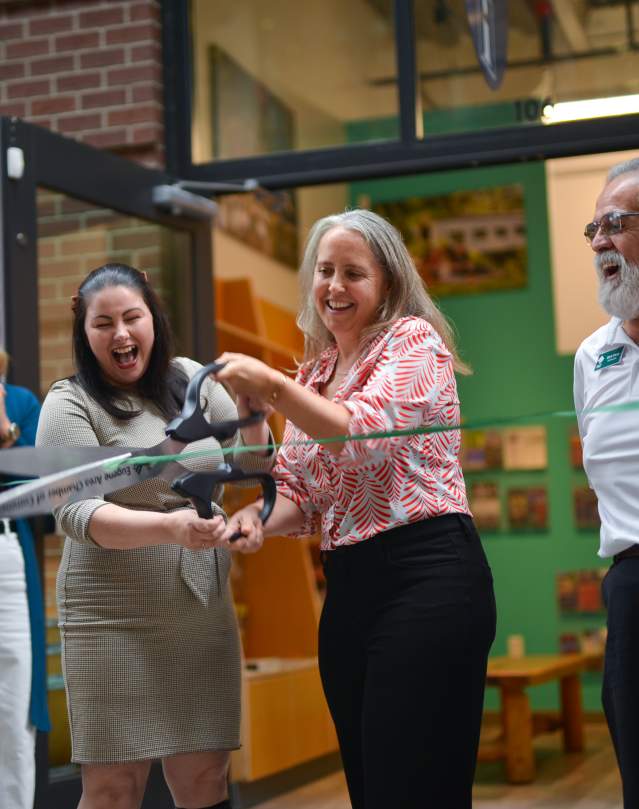 Travel Lane County's CEO cuts a green ribbon with giant scissors in front of the new Eugene, Cascades & Coast Visitor Center in Market Alley.