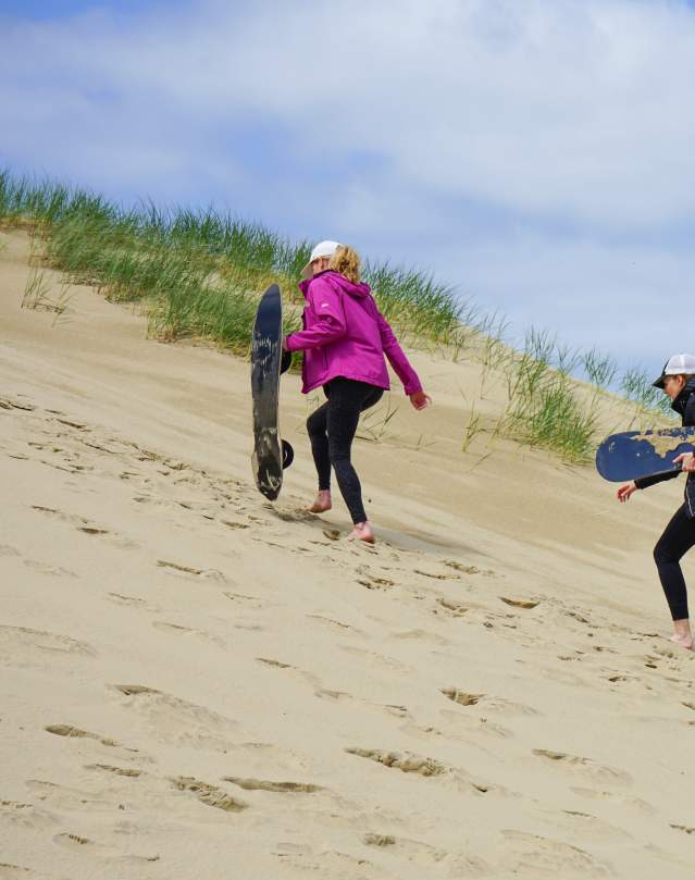 Three women with sandboards hike up a dune.