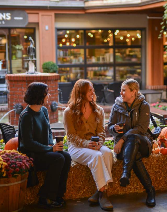 Three women enjoy coffee drinks sitting on haybales with pumpkins in the market courtyard.