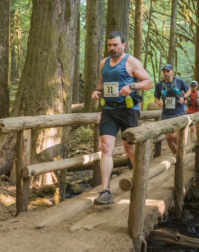 Three people run on a forested trail over a wooden footbridge.