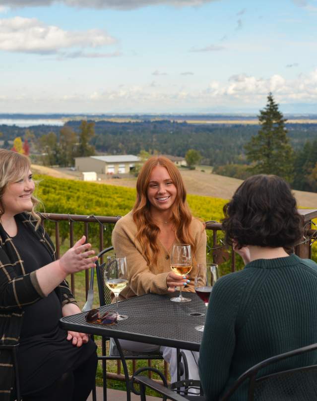 Four women taste wine at a patio table overlooking the Willamette Valley and vineyards.