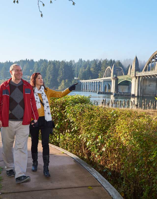 A couple walks near the Siuslaw River in Historic Old Town Florence with the beautiful highway 101 bridge in the background.