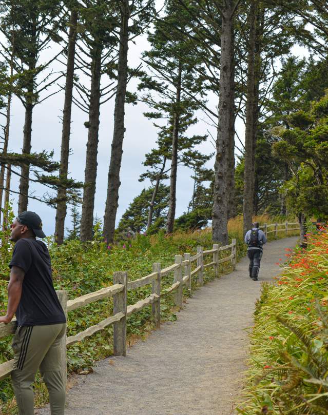 One man stands looking out over the ocean on a hiking trail. In the distance behind him, another man hikes up the hill.