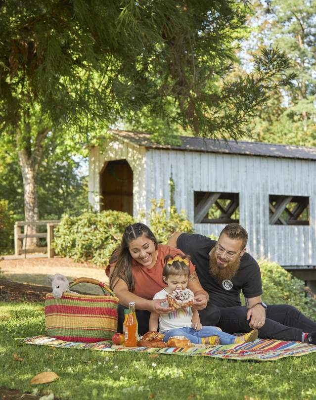 A family of three enjoys a picnic of pastries at a park with a white covered bridge in the background.