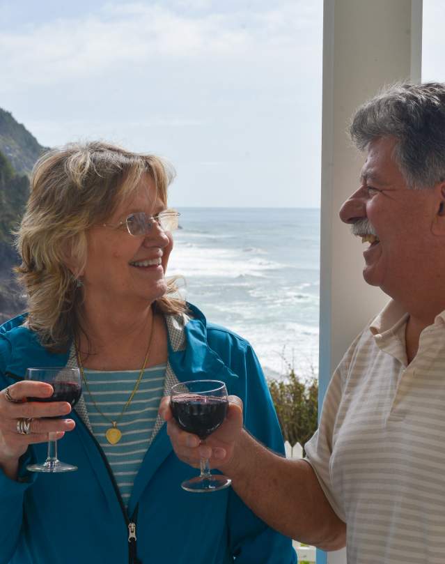 A couple drinking red wine is smiling with the ocean behind them.