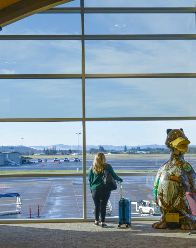 A woman with a rolling carry on suitcase stands in front of large windows at the airport looking out over the tarmac.