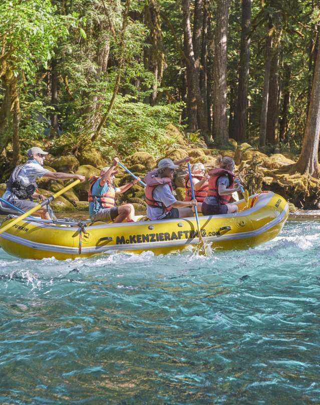 A raft full of smiling people paddles down a bright turquoise colored river surrounded by lush forest.