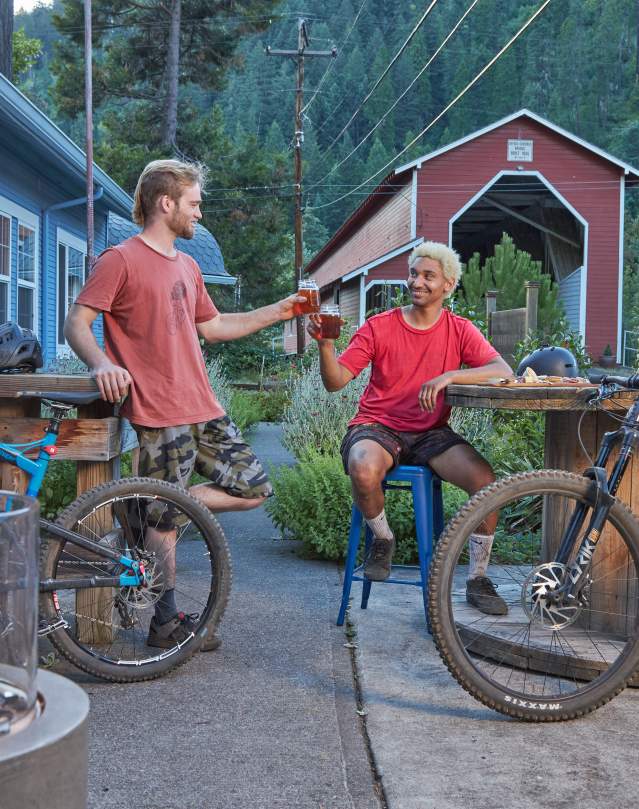 Two mountain bikers with their bikes nearby sit and cheers with their beers on a patio at Westfir Lodge in the evening. There is a fire in a firepit in the foreground and the red Office Covered Bridge in the background