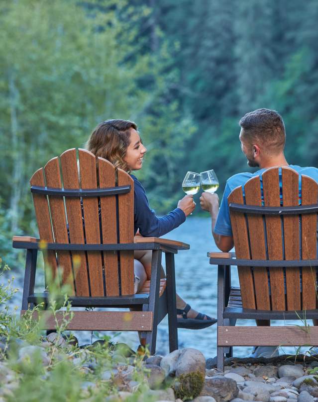 A couple sits in Adirondack chairs facing away from the camera beside the Mckenzie River in the evening. They are looking at each other and doing a cheers with their glasses of white wine.
